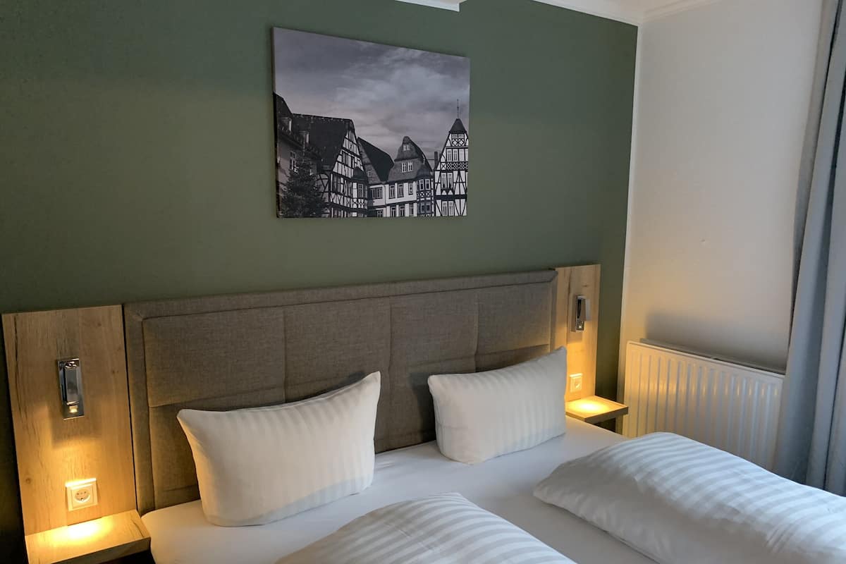 A hotel room with a large bed, two bedside tables, a headboard, and a painting of a European town on the wall.