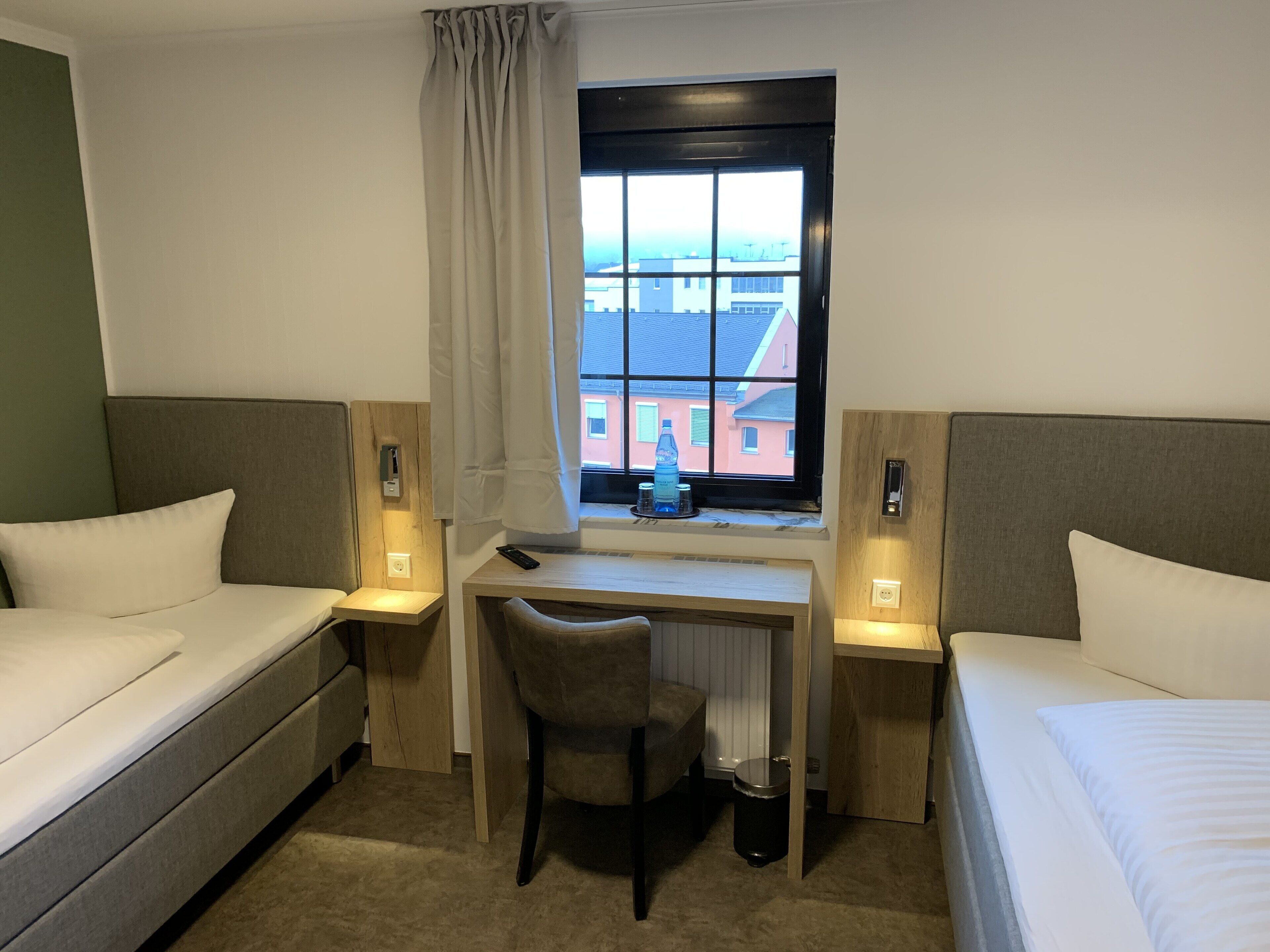 Ein Hotelzimmer mit zwei Einzelbetten, einem Schreibtisch mit Stuhl, einem Fenster mit Blick auf Gebäude und einer kleinen Mülltonne.
