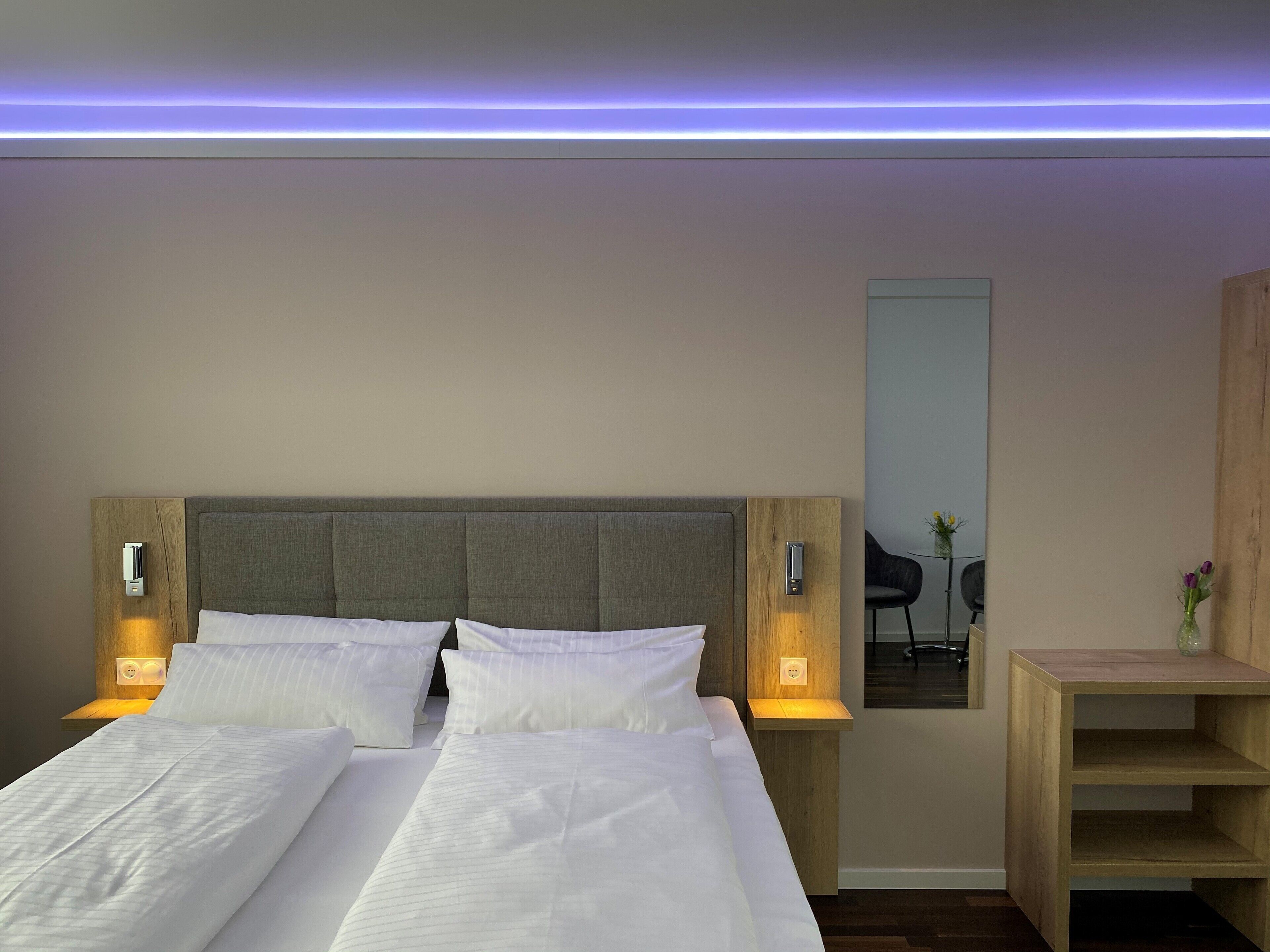 A hotel room with a double bed, two bedside tables with lamps, a wooden headboard, and a small wooden shelf.