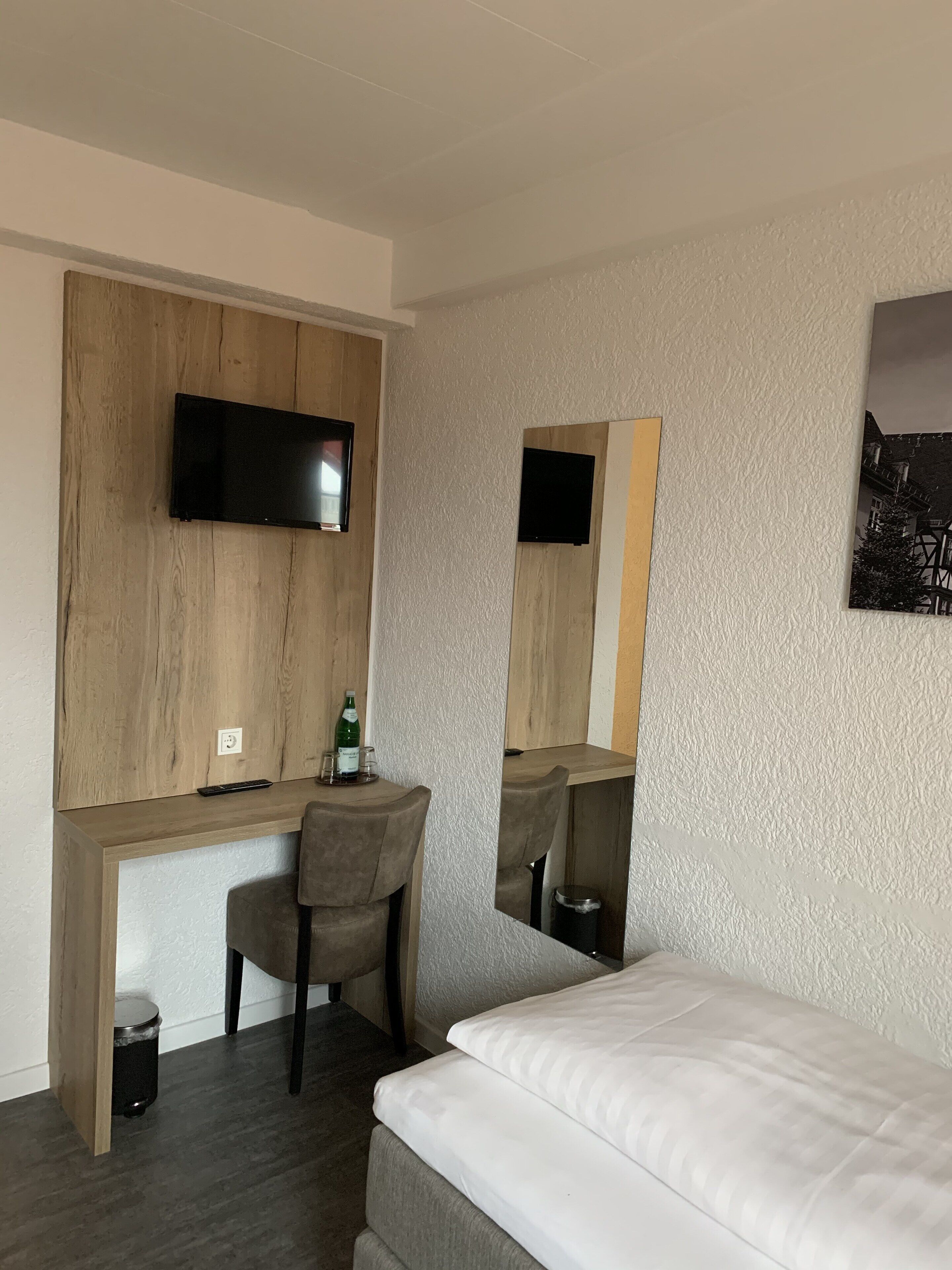A hotel room with a bed, a desk with a chair, a television mounted on a wooden panel, and a trash bin.