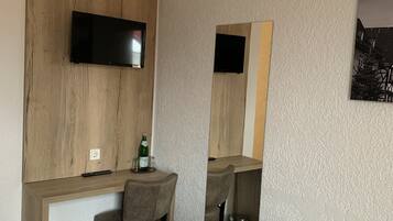 A hotel room with a bed, a desk with a chair, a television mounted on a wooden panel, and a trash bin.