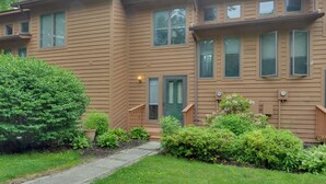 A two-story apartment building with a green door, a small front porch, and a well-maintained garden with various shrubs and a large tree. - Oakland Vacation Rental < 1 Mi to Deep Creek Lake! (Oakland)