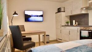 A compact hotel room with a bed, a desk, a chair, a television, and a kitchenette.
