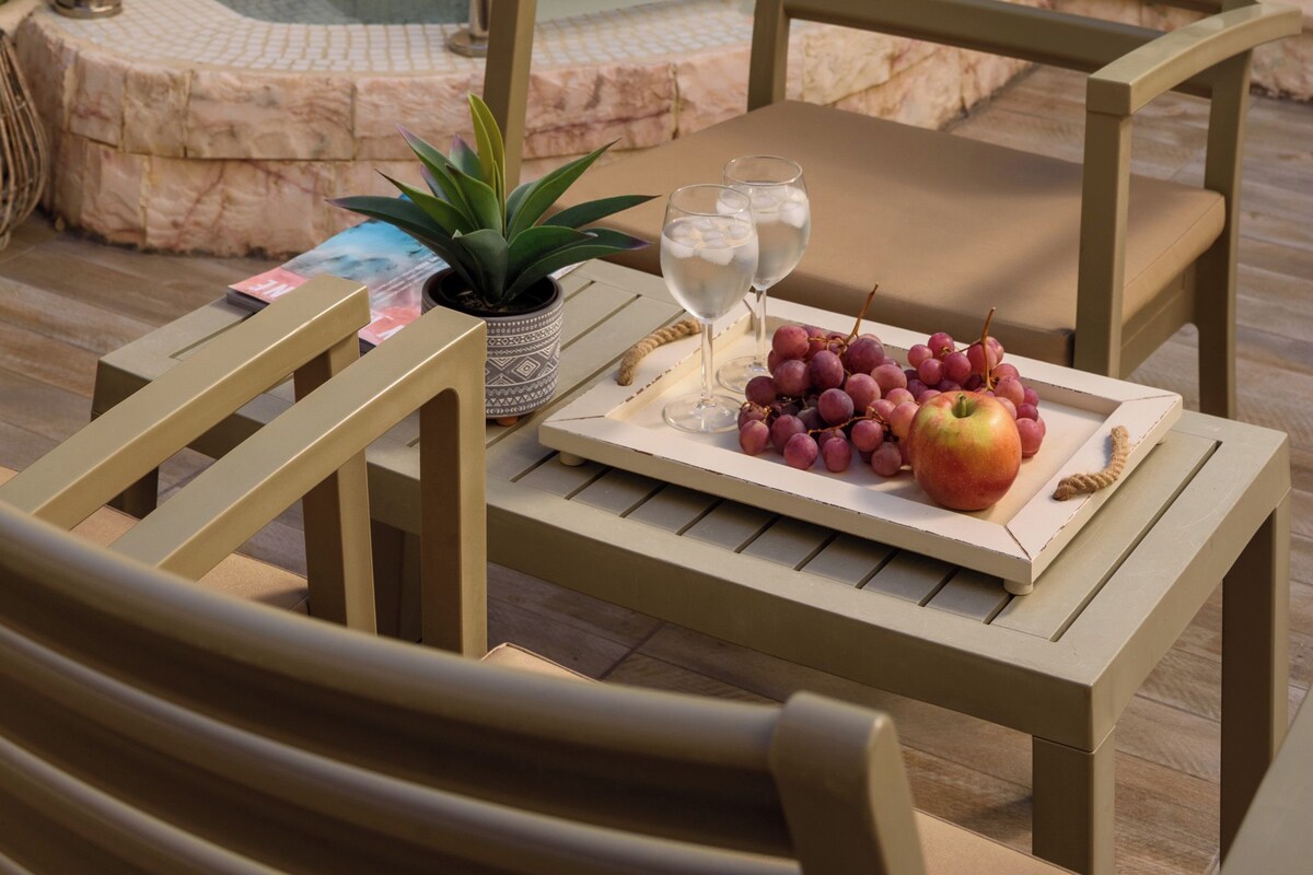 A stone wall with a built-in hot tub, a wooden chair, a table with a tray of fruit and wine glasses, and a small potted plant.