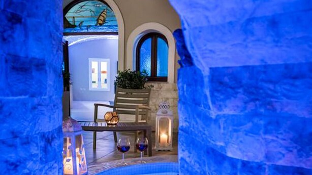 An indoor swimming pool with blue lighting, stone walls, and a seating area with a table and chairs. - Suite Home Santorini (Santorini)