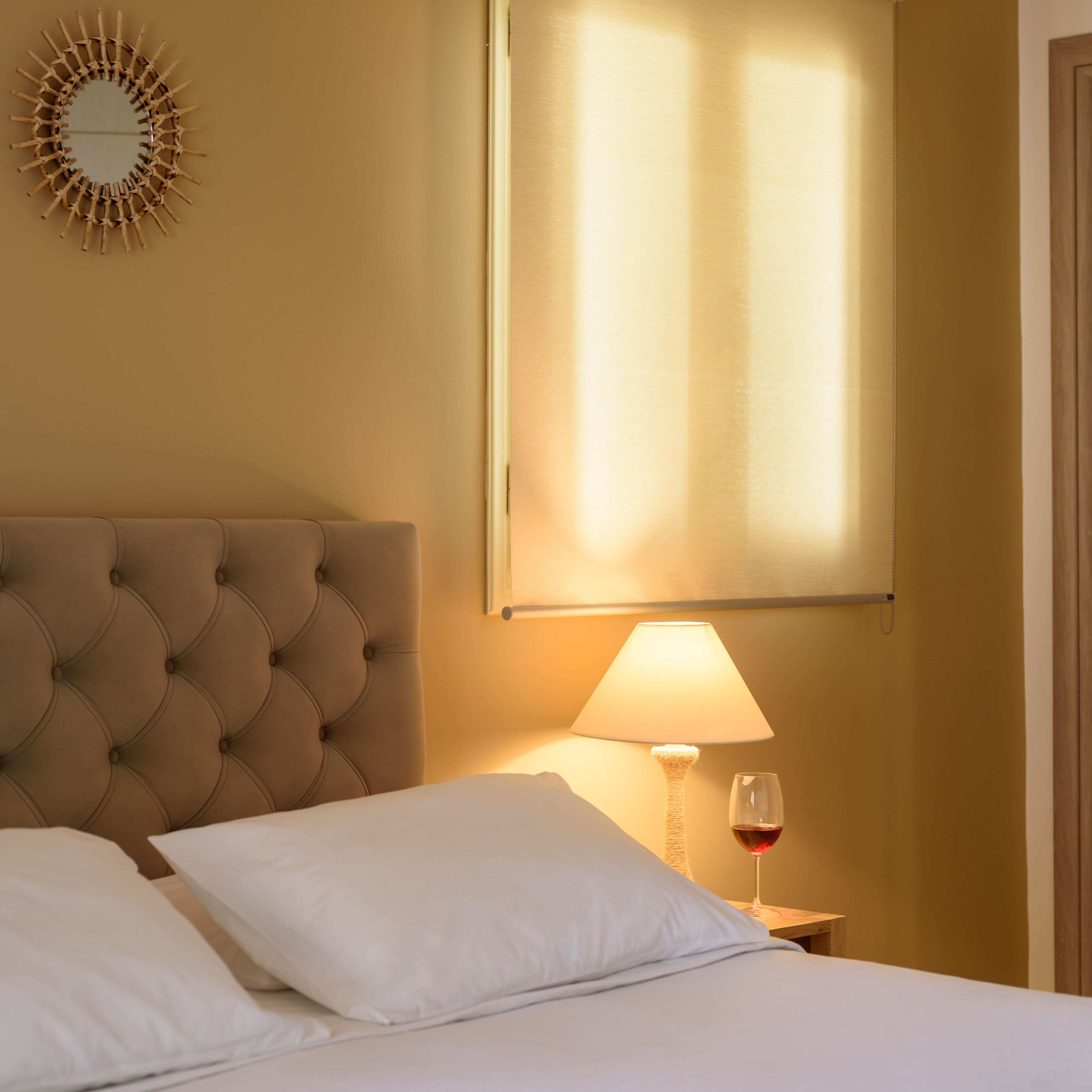 A hotel room with a tufted headboard, a bed with white linens, a bedside table with a lamp and a wine glass, and a wall-mounted mirror.