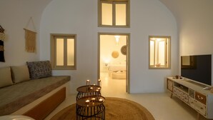 A cozy living room with a sofa, a TV, and a small table with candles. There are two windows and a door leading to another room. - Suite Home Santorini (Santorini)