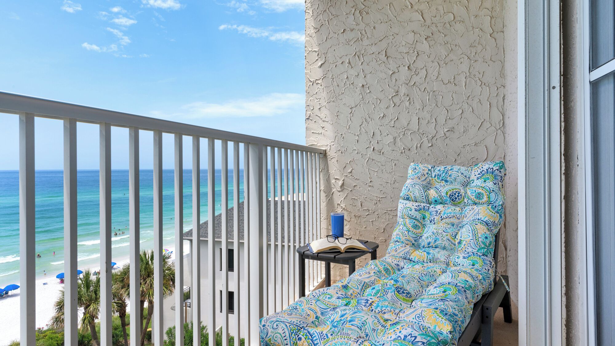 A balcony with a view of the beach, a patterned chair, and a small table with a book and glasses.