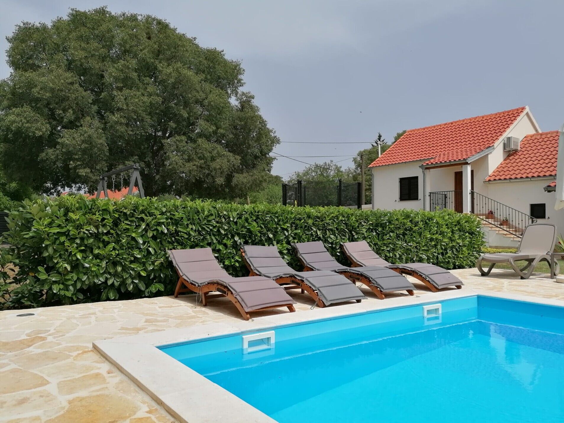 Une piscine avec des chaises longues, une haie et un bâtiment doté d’un toit rouge.