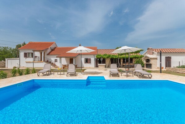 A swimming pool with lounge chairs and umbrellas, surrounded by white buildings with red-tiled roofs. - Villa Vitis (Promina)