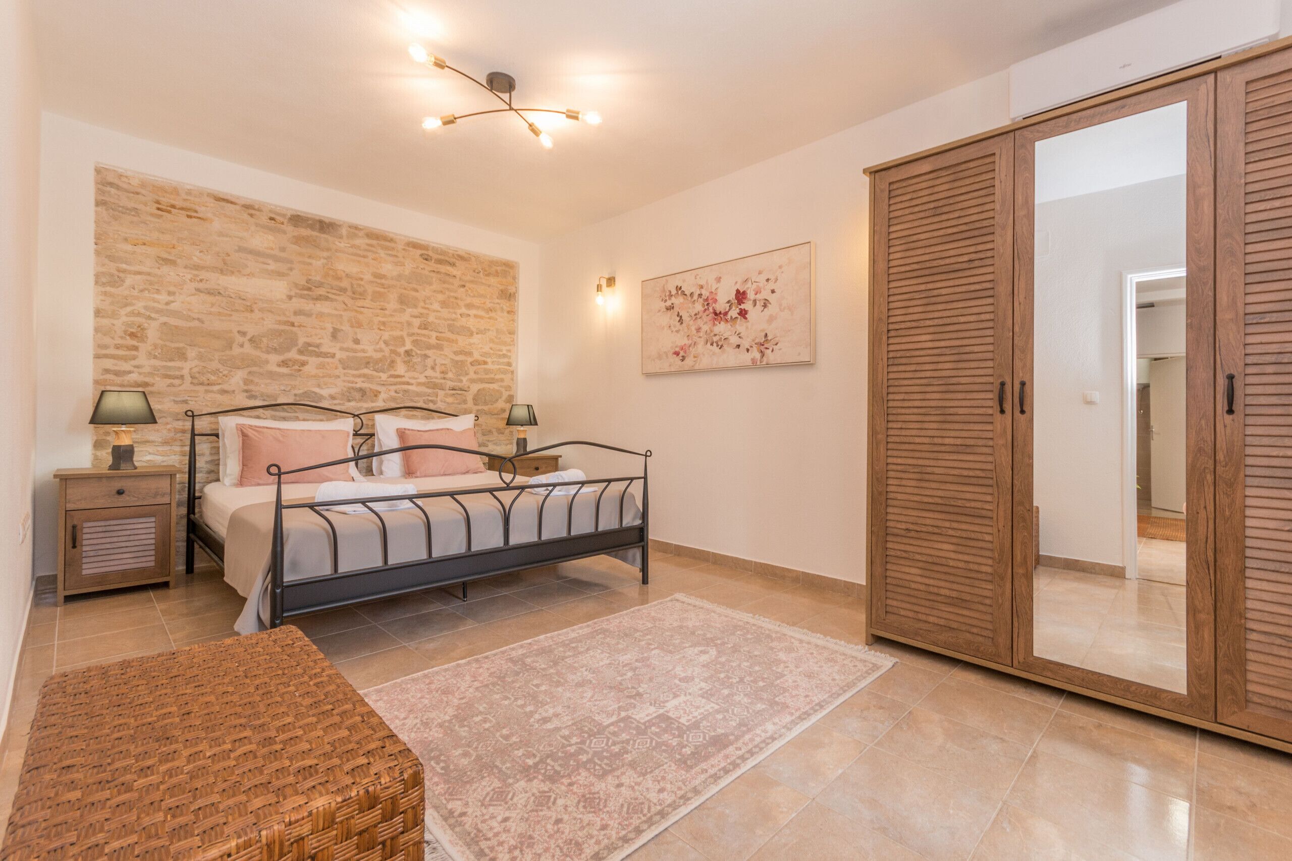 Une chambre avec un mur en pierre, un lit avec une tête de lit, une armoire en bois, une table de chevet et une lampe sur pied.