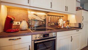 Une cuisine avec des armoires blanches, un four en acier inoxydable, une machine à café rouge et un pare-feu décoré d’un motif représentant un paysage.