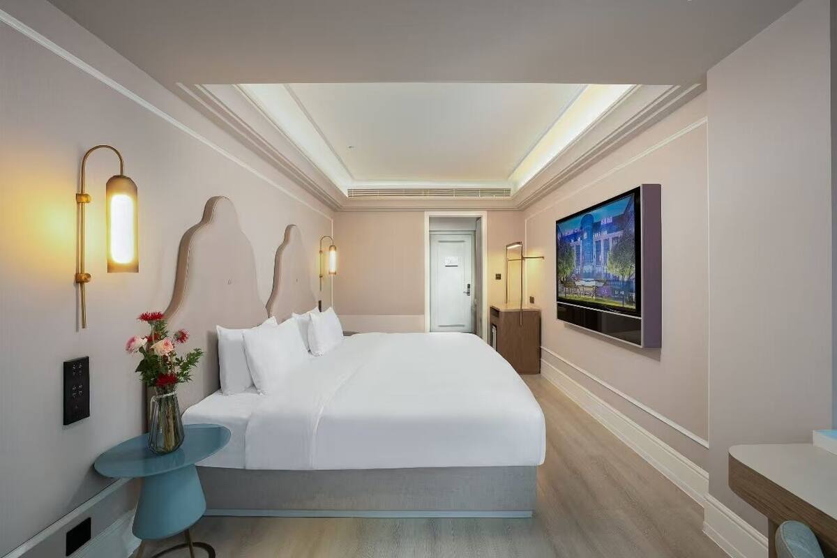 Ein modernes Hotelzimmer mit einem großen Bett, einem Flachbildfernseher, einem kleinen Beistelltisch und einer Vase mit Blumen.