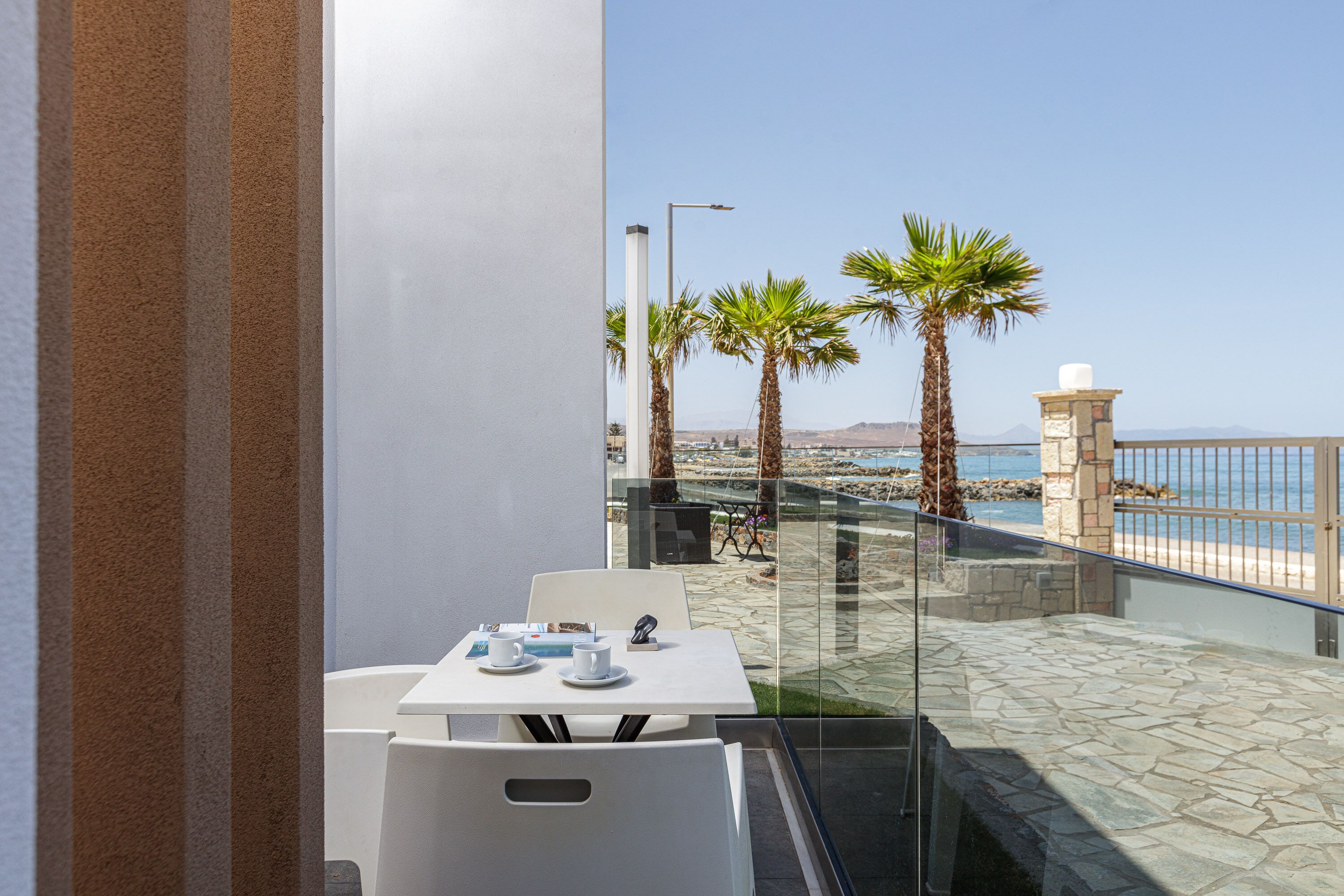 Terraço com mesa e cadeiras, vista para uma praia com palmeiras e céu claro.