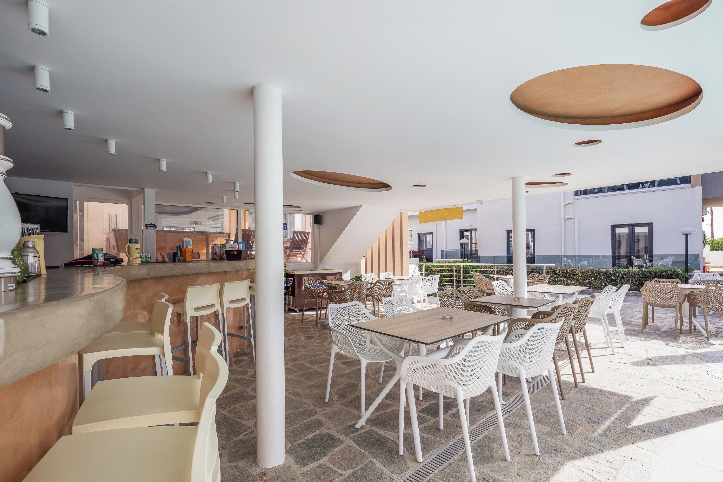 Un área de comedor exterior moderna con sillas y mesas de mimbre blanco, una barra y vistas del interior del edificio.