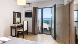 A hotel room with a bed, a desk with a chair, a TV, a potted plant, a sliding glass door to a balcony with a view of the sea, and an air conditioning unit.