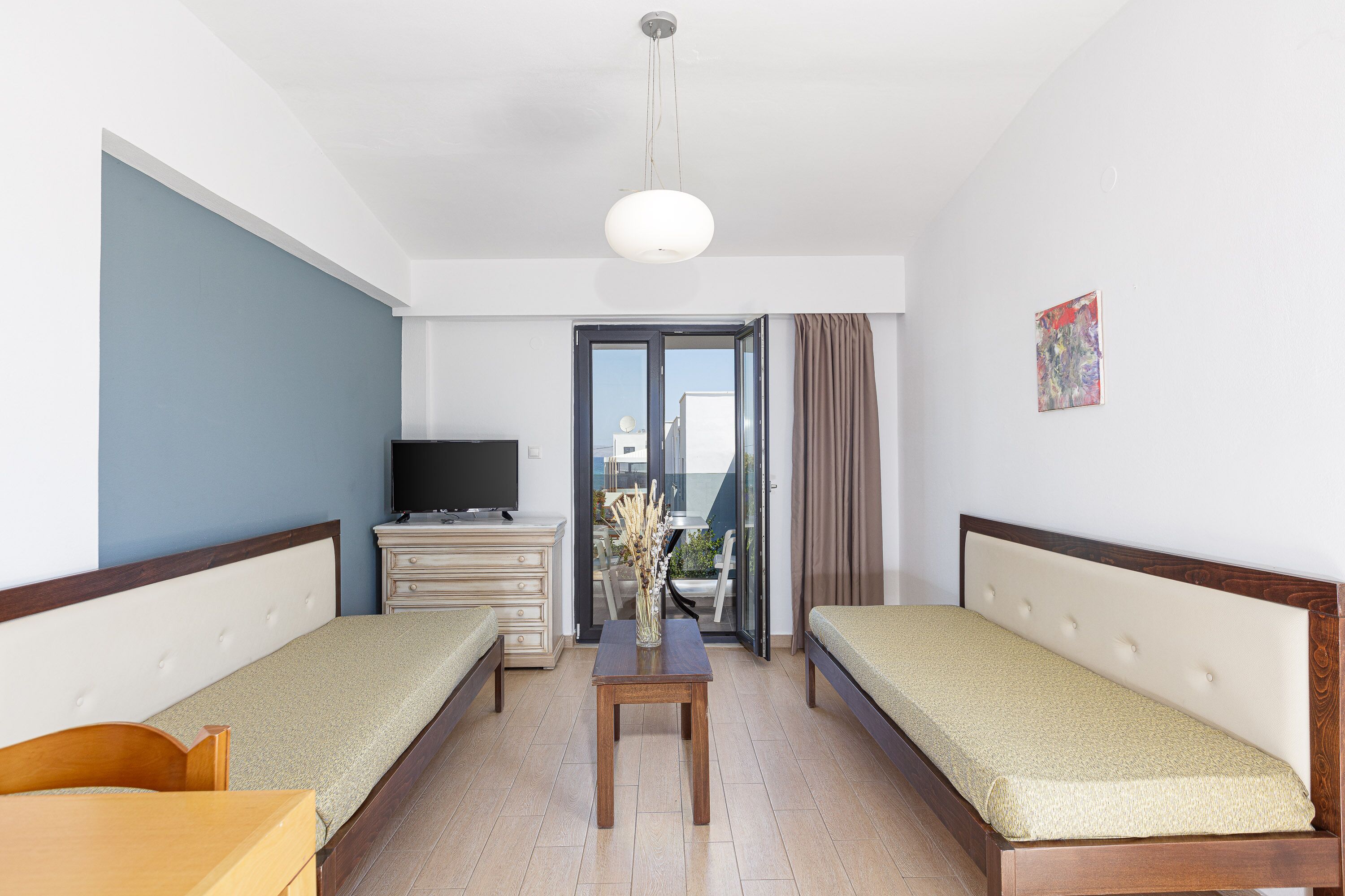 A room with two single beds, a small table, a TV, and a view of the outside.