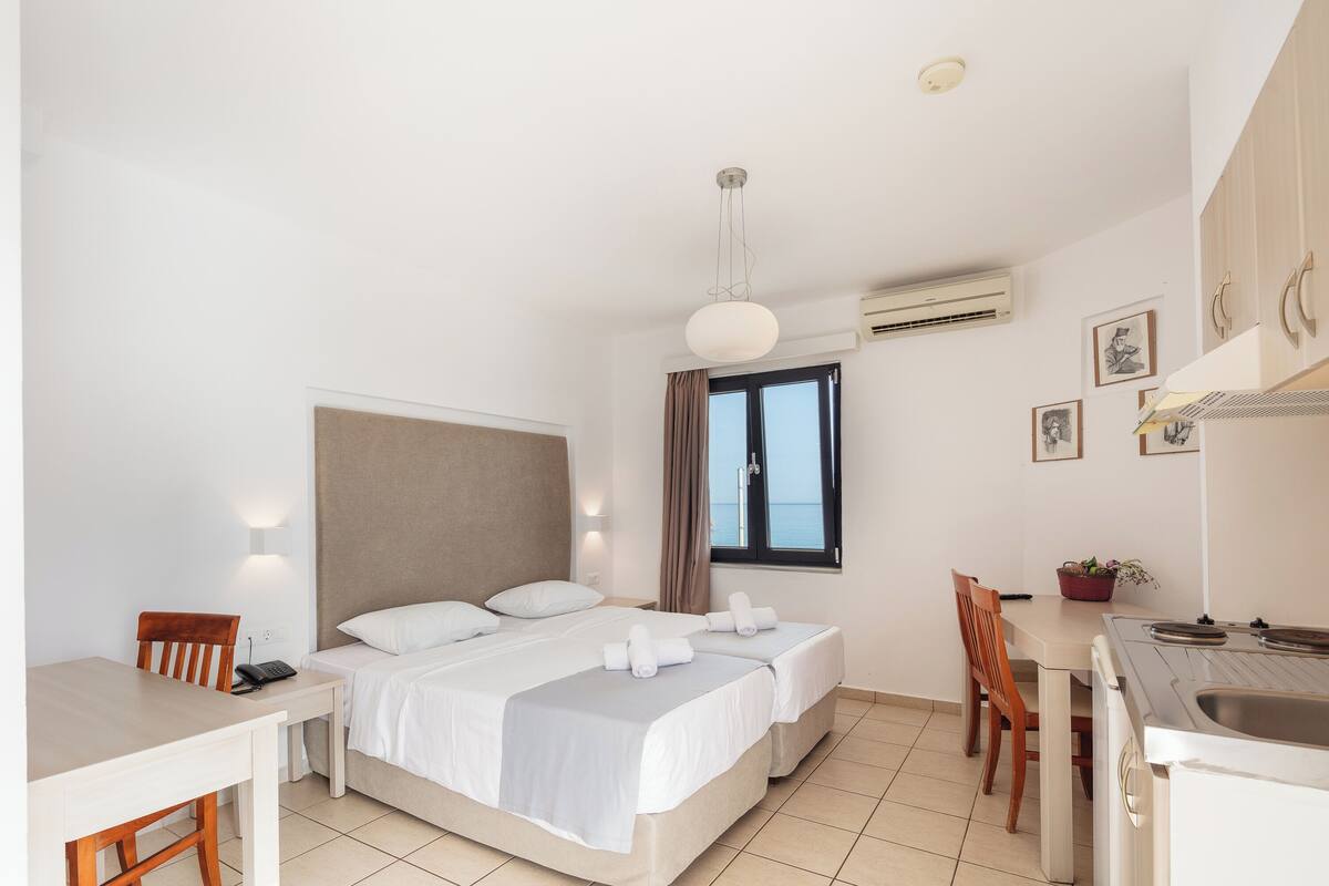 A hotel room with a bed, a desk, a chair, a small table, a window with curtains, and a sink.