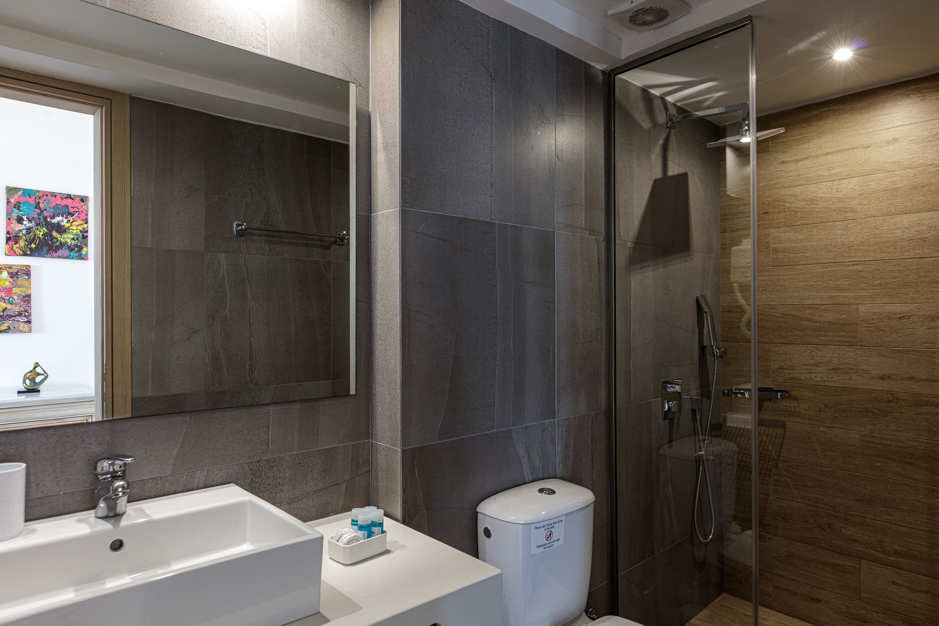 A modern bathroom with a large mirror, a white sink, a glass-enclosed shower, and a wooden wall.