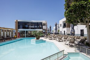 A pool area with lounge chairs, a Coca-Cola cooler, and a modern building with a glass facade. - Golden Bay Boutique Hotel & Bungalows (Hersonissos)