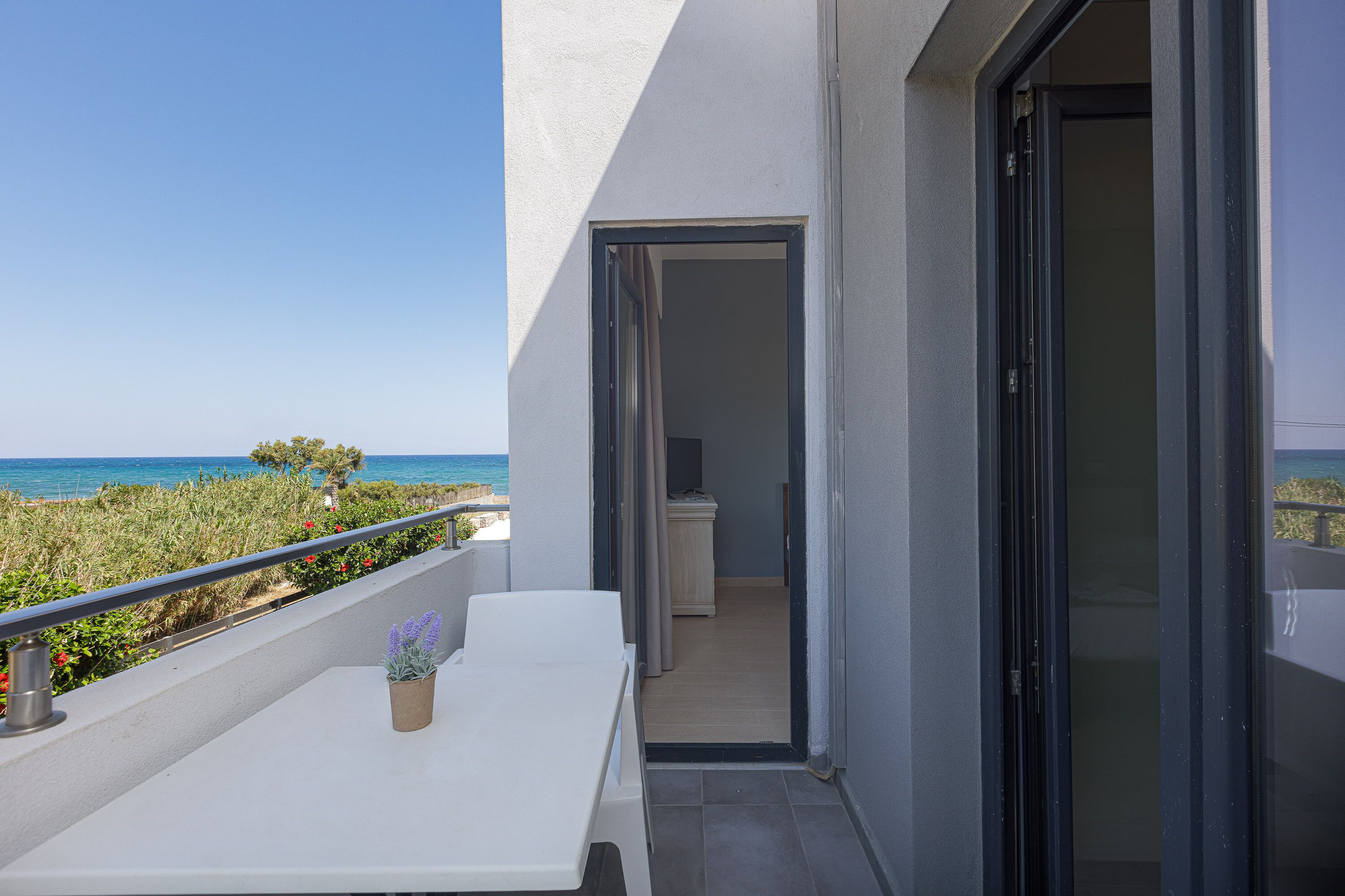 A balcony with a white table and a potted plant, overlooking a coastal area with a clear sky.