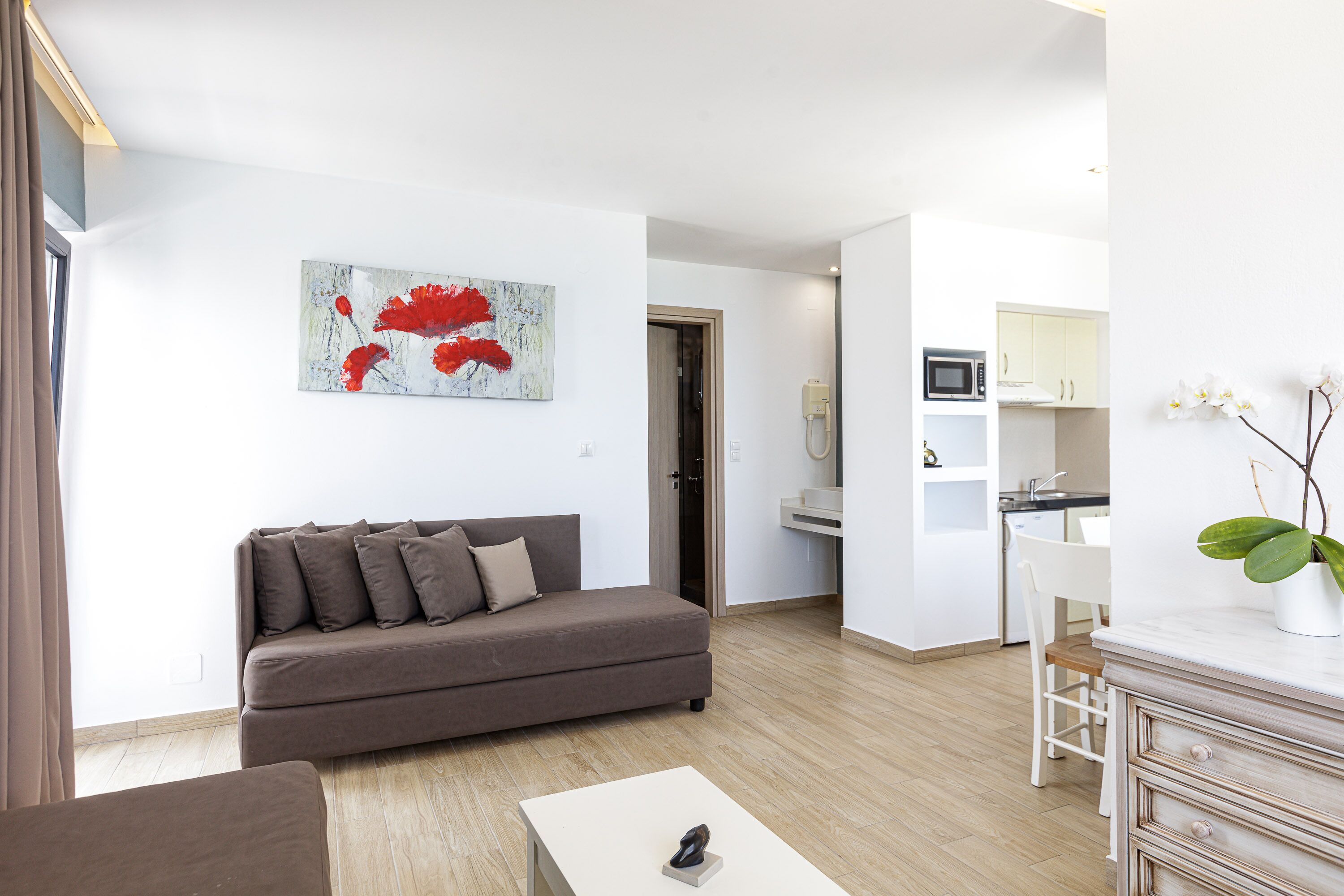 Een moderne woonkamer met een bruine bank, een witte salontafel en een keuken met witte kastjes en een magnetron.