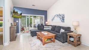 A living room with a sofa, coffee table, and side table, a TV mounted on the wall, and a sliding glass door leading to an outdoor area. - TB 94B Dog friendly Garden home in the peaceful True Blue community. (Pawleys island)