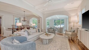 Un salon spacieux avec un grand canapé, une table basse et un téléviseur. On y trouve plusieurs options d’assises, notamment un fauteuil et un divan. La pièce est dotée d’un haut plafond avec un ventilateur et de grandes fenêtres qui laissent entrer la lumière naturelle.