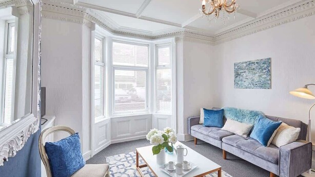 A room with a white ceiling, a chandelier, a grey sofa with blue and white cushions, a white coffee table, and a window with white shutters. - Host & Stay | Ocean Breeze (Scarborough)