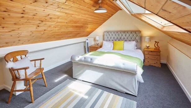 A bedroom with a sloped ceiling, a bed with a tufted headboard, a wooden chair, and a bedside table with a lamp.