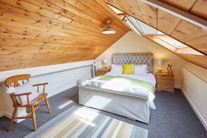 A bedroom with a sloped ceiling, a bed with a tufted headboard, a wooden chair, and a bedside table with a lamp.