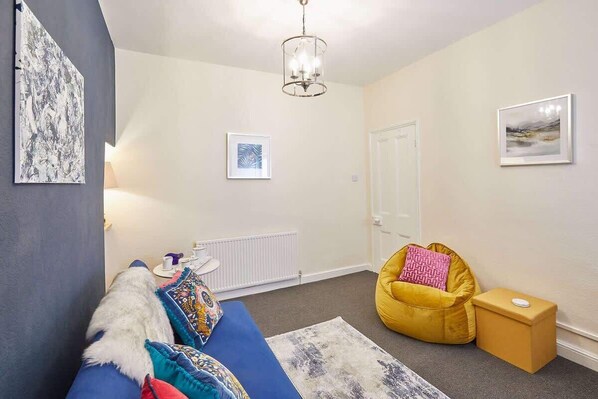 A cozy living room with a blue sofa, a yellow bean bag, a small table with a lamp, and a radiator.