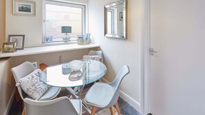 A dining area with a round glass table, white chairs, a small side table with a lamp, and framed pictures on the wall.