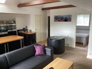 A modern living room with a sofa, a coffee table, a round black ottoman, a wooden dining table, and a kitchen area with a microwave and oven.