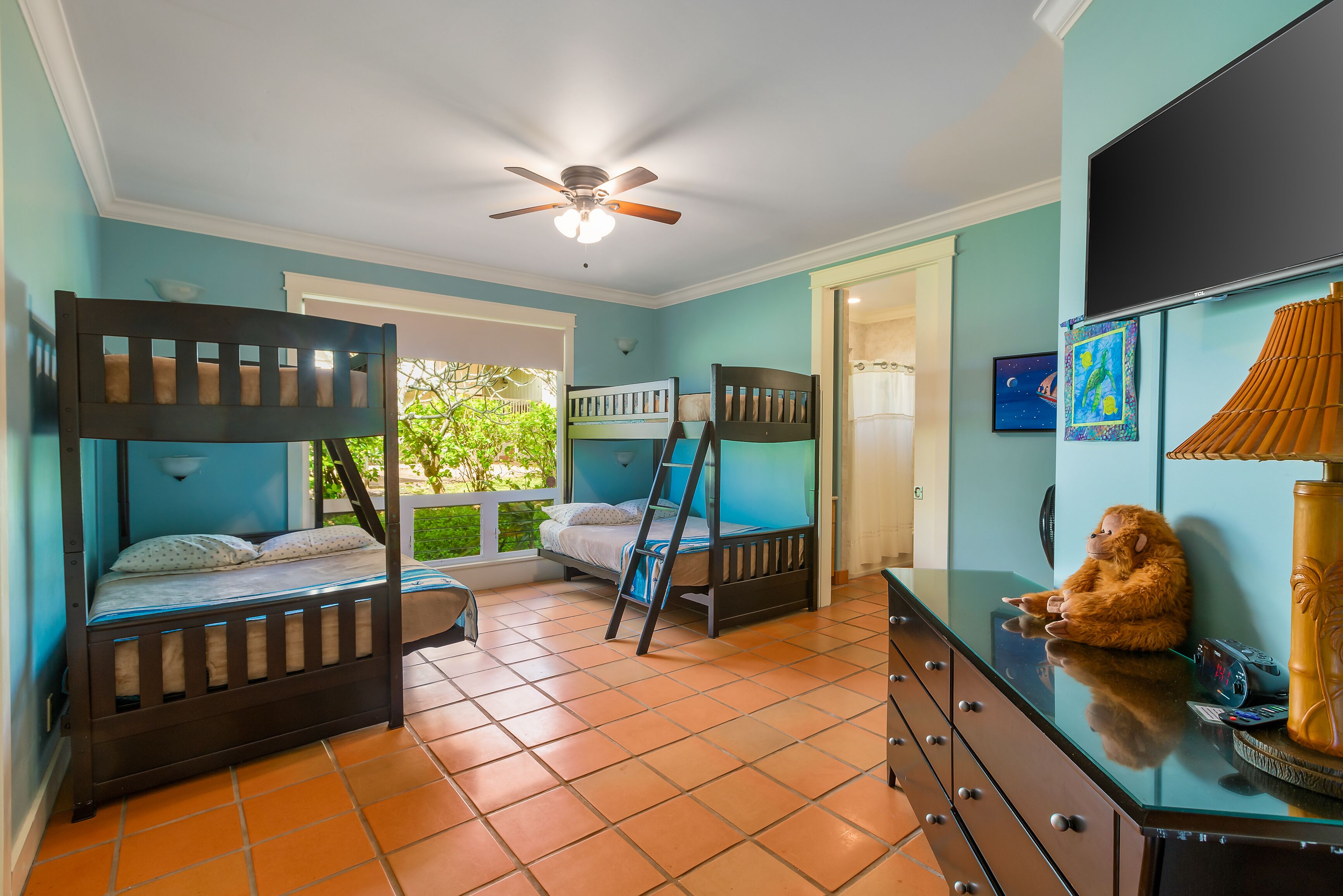 A bedroom with two bunk beds, a ceiling fan, a dresser, and a window with a view of greenery.
