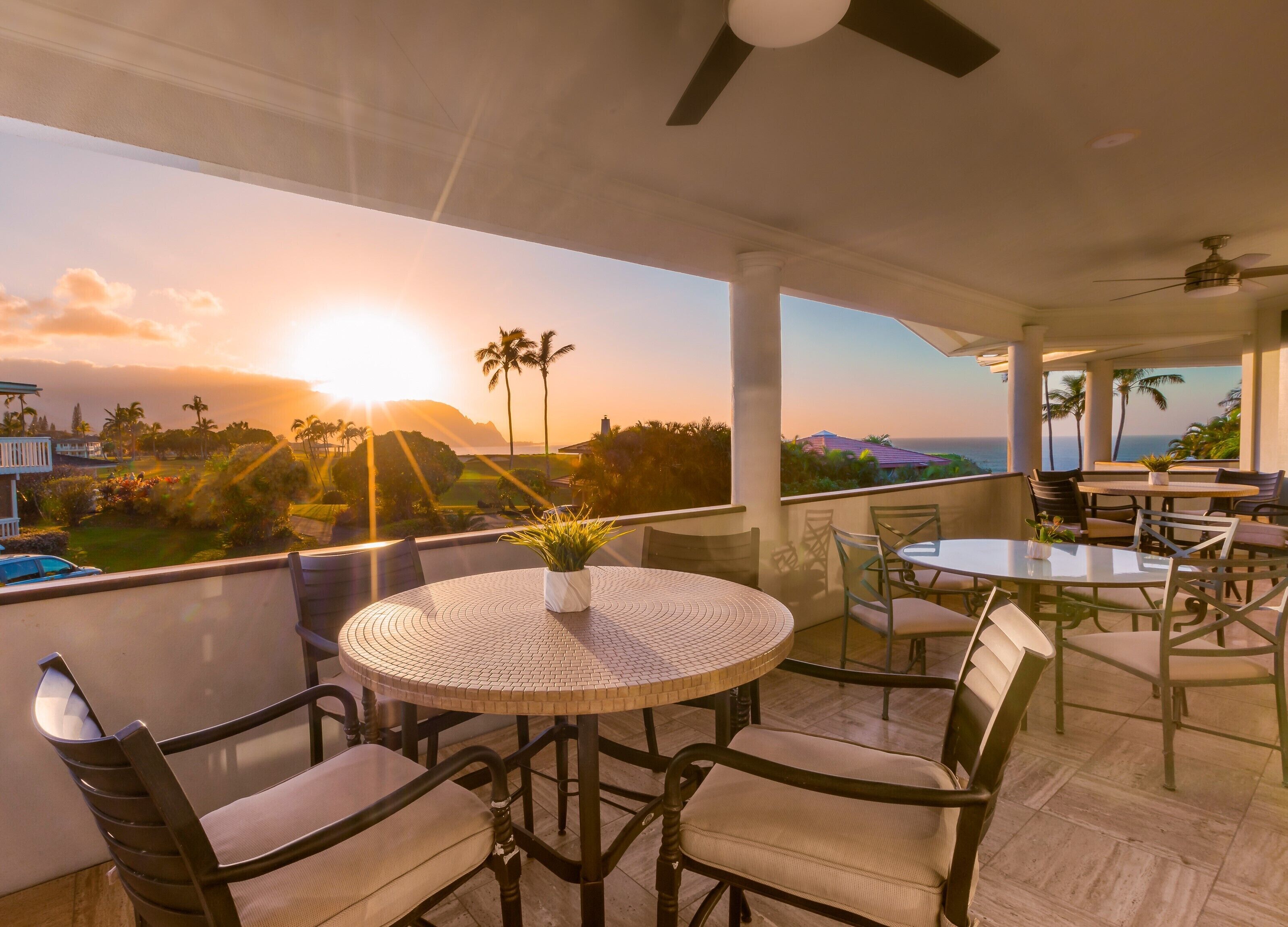 A balcony with round tables and chairs, overlooking a scenic view of the ocean and palm trees during sunset.