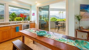 A kitchen with a wooden table, a window with a view of the outdoors, and a sliding glass door leading to a patio. - Hawaii Life Rentals Presents Hale Ohana Nui (Princeville)
