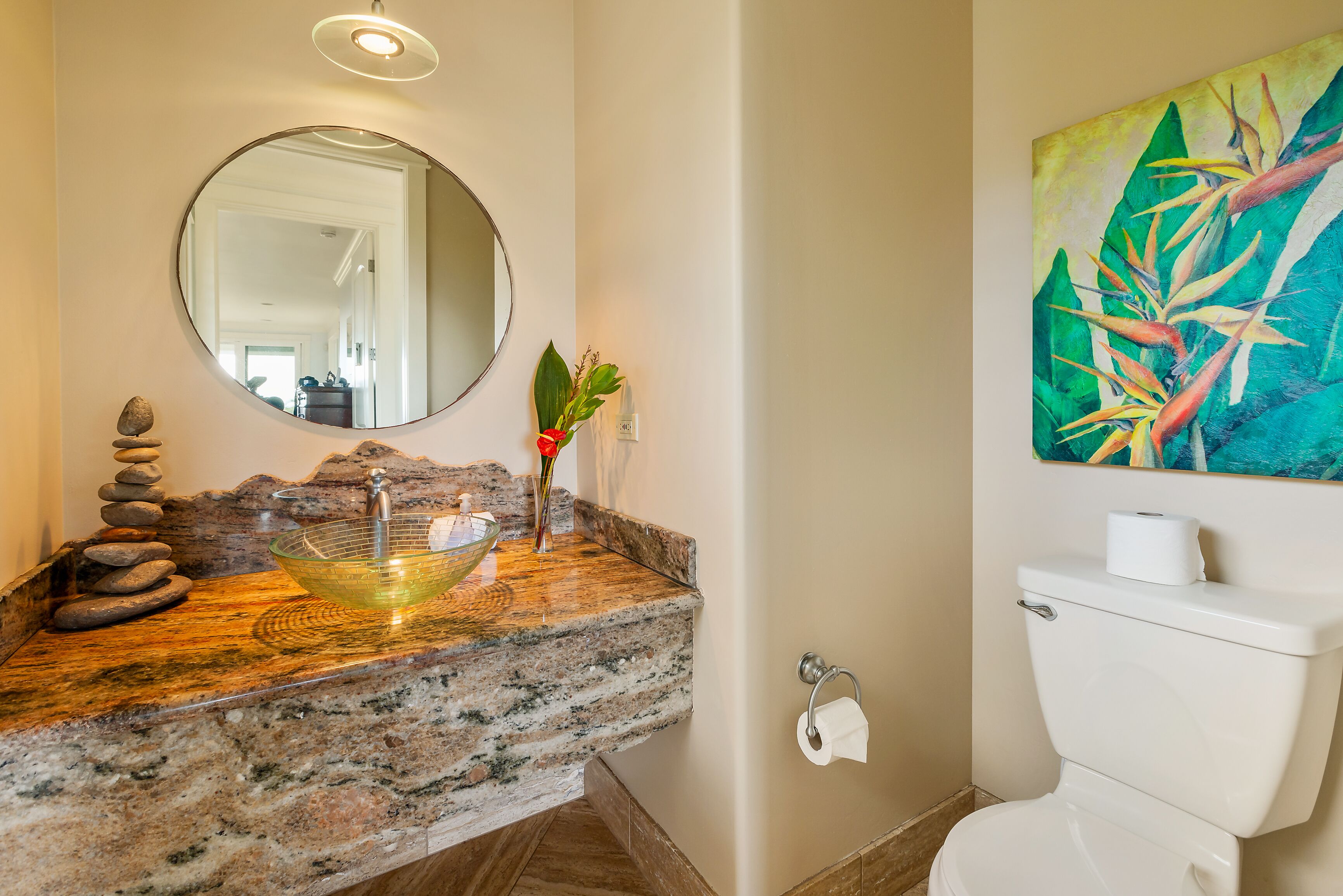 Un bagno con un piano di lavoro in granito, uno specchio rotondo, un lavabo in vetro, un water e un quadro con foglie tropicali.