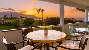 A round table with a woven top, surrounded by chairs, overlooking a scenic view of a sunset over a coastal landscape with palm trees and mountains. - Hawaii Life Rentals Presents Hale Ohana Nui (Princeville)