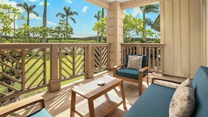 Un espacio de estar al aire libre techado con una mesa y sillas de madera, un sofá con cojines y vistas a un jardín tropical con palmeras.