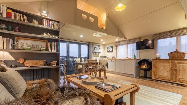 A spacious living area with a high ceiling, wooden furniture, a fireplace, and a large window overlooking a cityscape. - The Bothy, Ringstead, Norfolk (Ringstead)