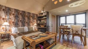 A bedroom with a large bed, a wooden side table, a bookshelf, a dining area with a white furry chair, and a view through large windows. - The Bothy, Ringstead, Norfolk (Ringstead)