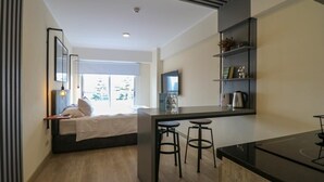 Un dormitorio moderno con cama, mesita de noche y ventana. Hay una kitchenette con estufa, taburetes de bar y televisor.