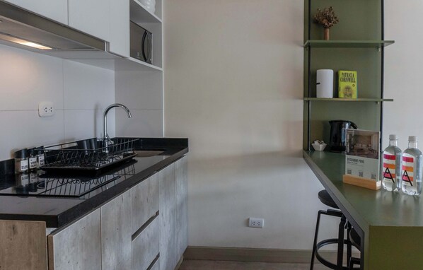 A modern kitchen with a black countertop, stainless steel sink, and a coffee machine. There are two bar stools and a shelf with a book and a vase.