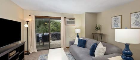 A living room with a flat-screen TV, grey sofa, blue cushions, a white coffee table, and a blue lamp.