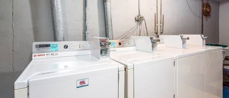 A row of white washers with visible brand labels, connected to a grey wall with metal pipes and electrical outlets.
