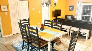 A dining area with a table set for four, surrounded by chairs. There is a sofa and a television in the background. - 20 Steps to the Beach  -SUMMER DISCOUNT- (Grand Cayman)