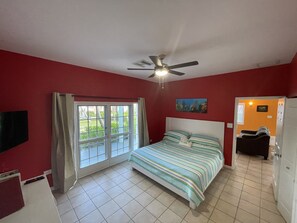 Un dormitorio con cama, ventilador de techo, ventana con cortinas y puerta que da a otra habitación.