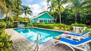 A swimming pool with lounge chairs, a lifeguard sign, and a house in the background. - 20 Steps to the Beach  -SUMMER DISCOUNT- (Grand Cayman)