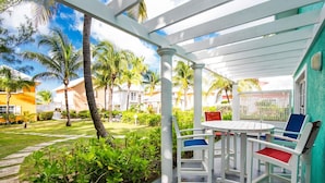 A covered outdoor seating area with a white table and chairs, surrounded by lush greenery and palm trees. - 20 Steps to the Beach  -SUMMER DISCOUNT- (Grand Cayman)